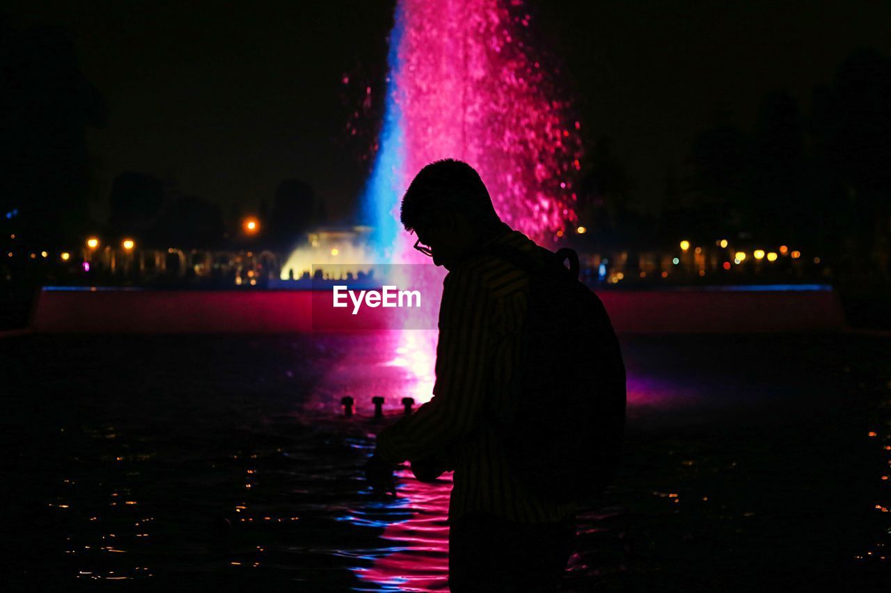 Side view of silhouette man standing against illuminated fountain at night