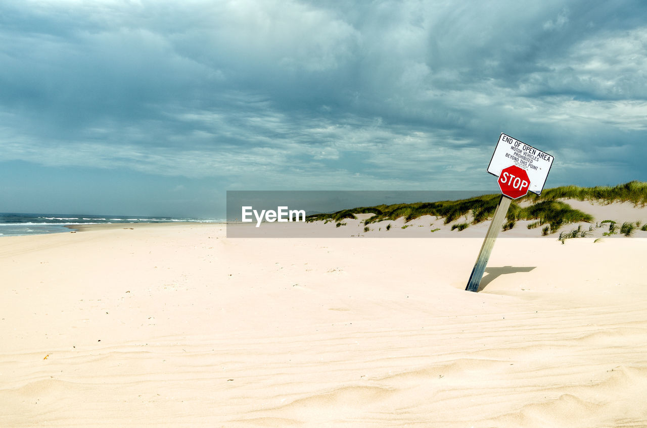 Stop sign on sand at beach against cloudy sky