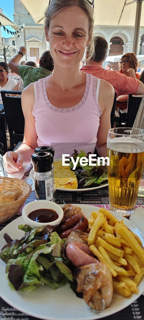 food and drink, food, women, adult, fast food, lunch, meal, smiling, dish, table, happiness, brunch, drink, female, vegetable, refreshment, person, restaurant, business, french fries, raw potato, two people, lifestyles, portrait, emotion, alcohol, glass, mature adult, holding, waist up, healthy eating, plate, enjoyment, cheerful, breakfast, sitting, cooking, leisure activity, meat, young adult, freshness, cuisine, front view, indoors, eating, togetherness, friendship, drinking glass, dinner