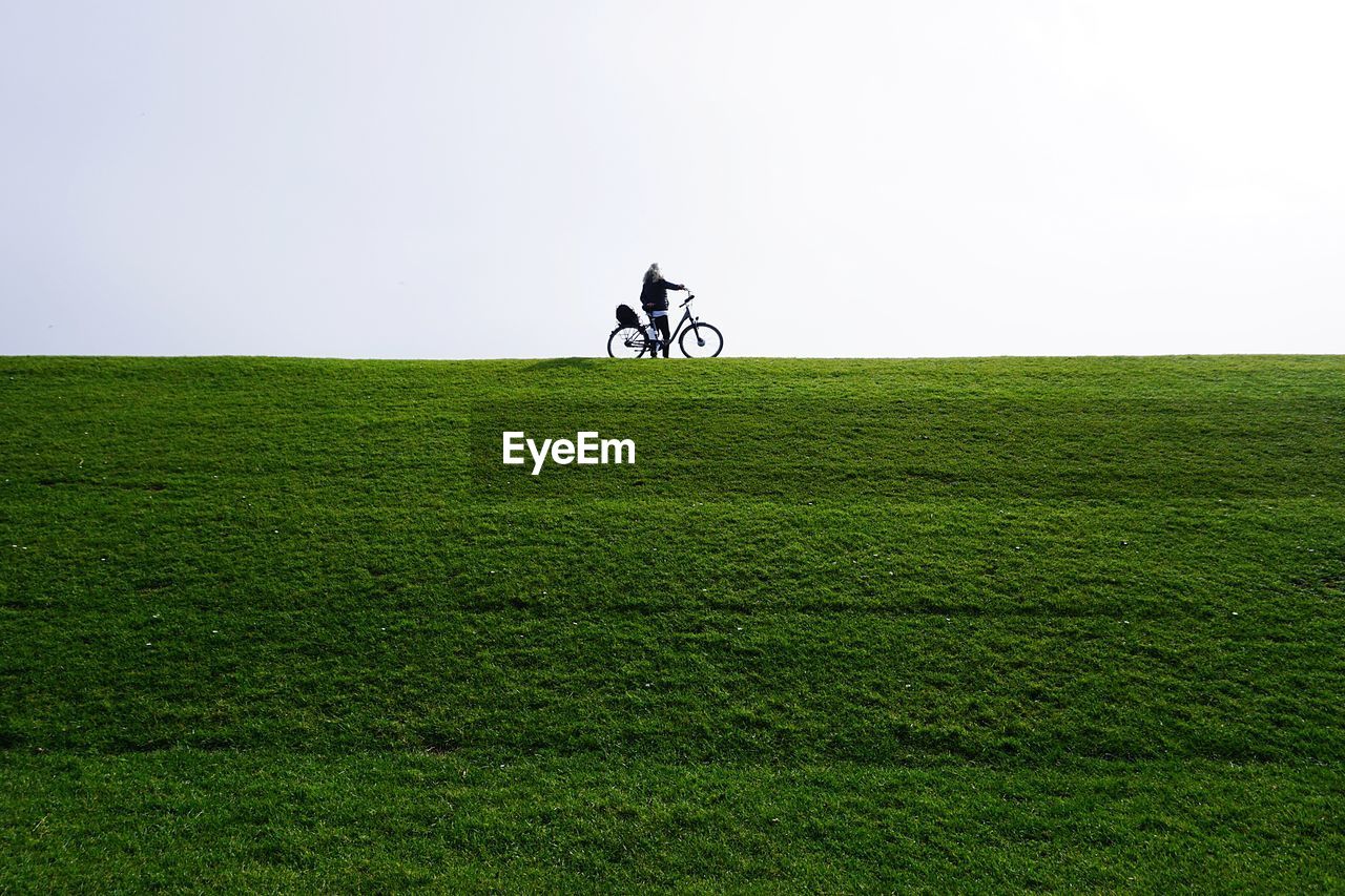 Distant view of woman with bicycle standing on grassy land against clear sky