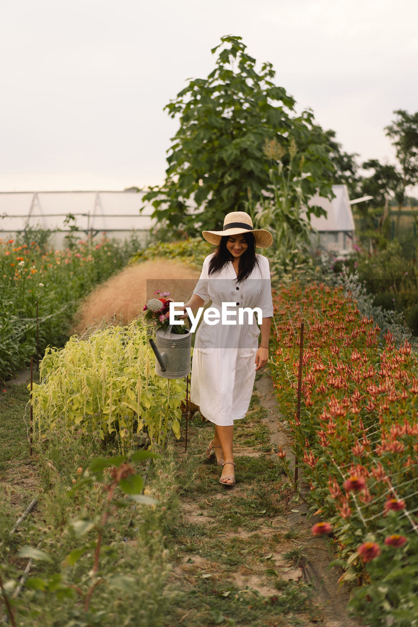 Beautiful brunette girl in a white dress and hat with watering can watering flowers