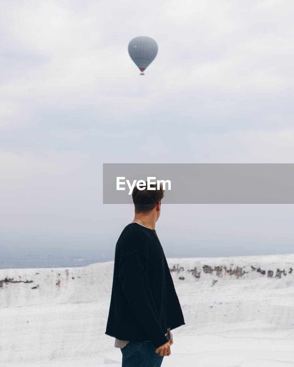 Traveling unrecognizable man in casual clothes looking away while standing on white hill of mineral formation against countryside at horizon and air balloon flying in gray sky in turkey