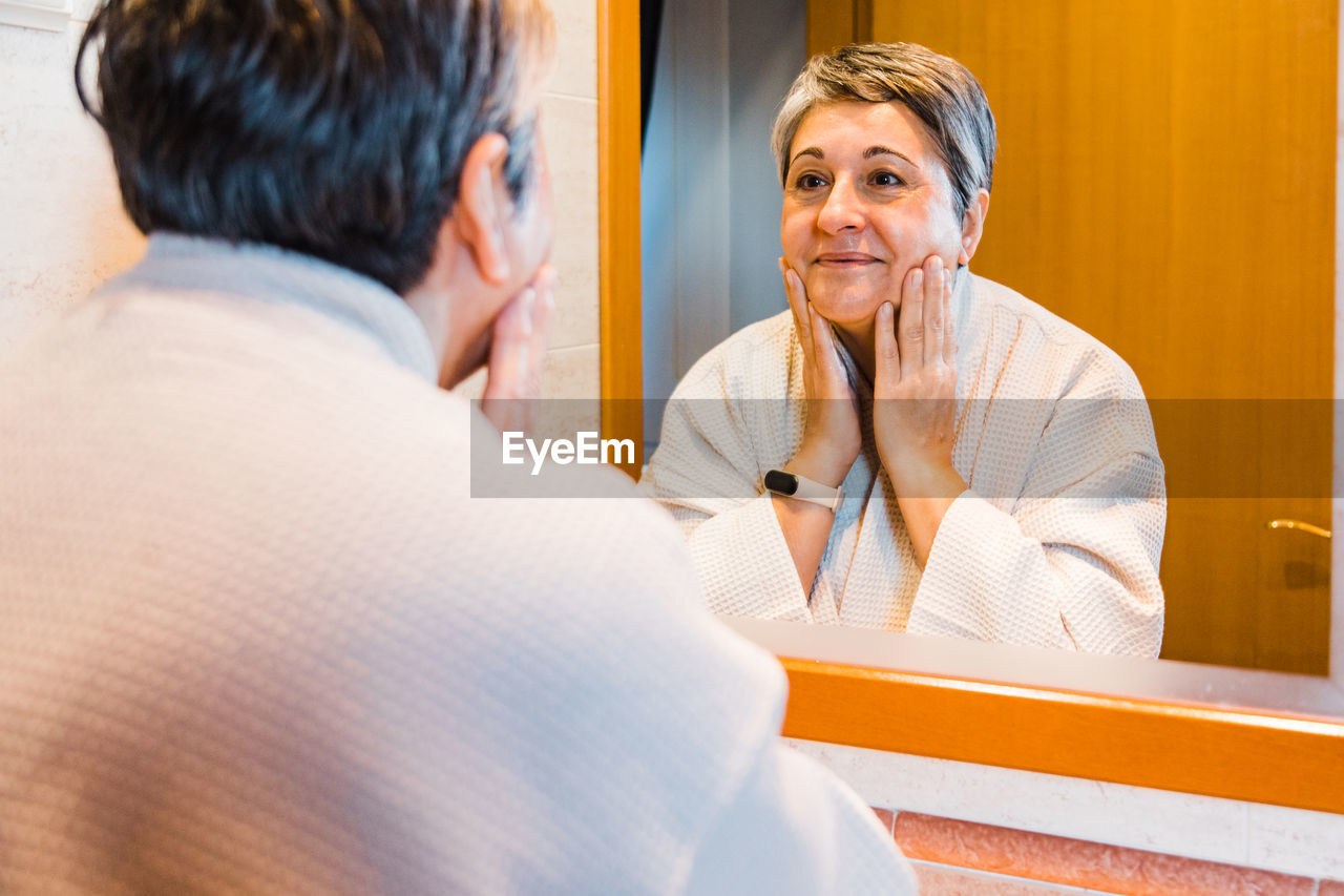 Middle aged female in white bathrobe touching face and looking into mirror during morning routine in light bathroom at home