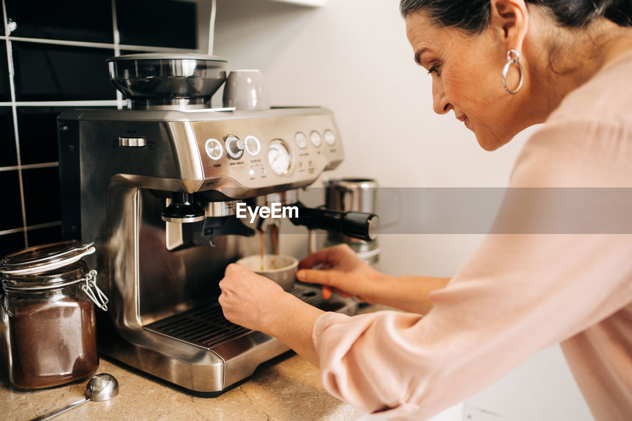 Side view of concentrated middle aged female preparing fresh coffee while using modern coffee maker at kitchen counter