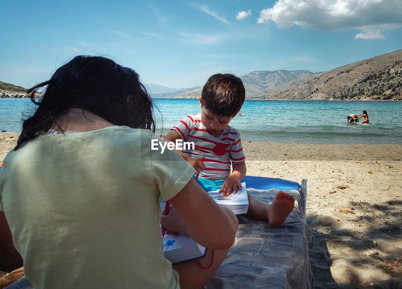 Siblings drawing while sitting on lounge chair at beach