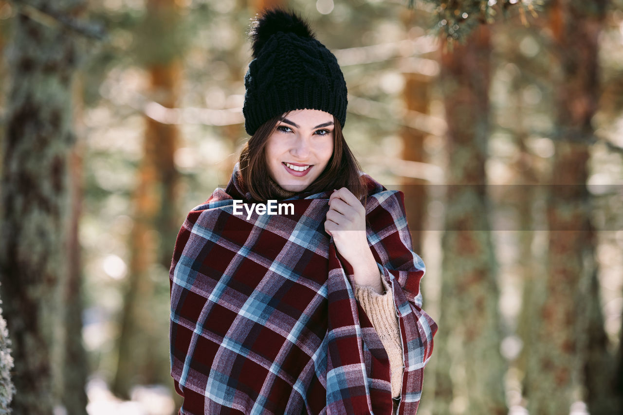 Content female wrapped in warm plaid standing in snowy woods and looking at camera