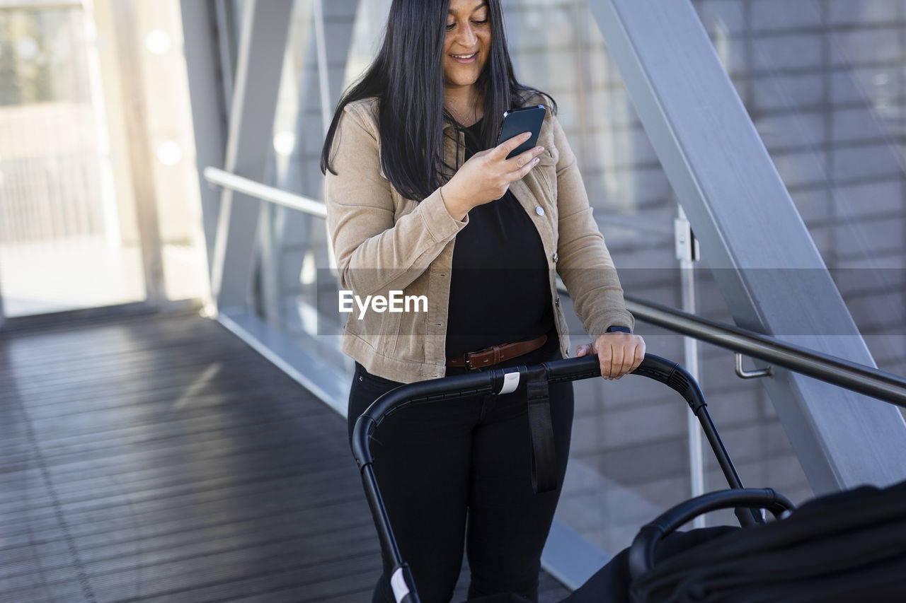 Smiling woman on parental leave with pram using cell phone