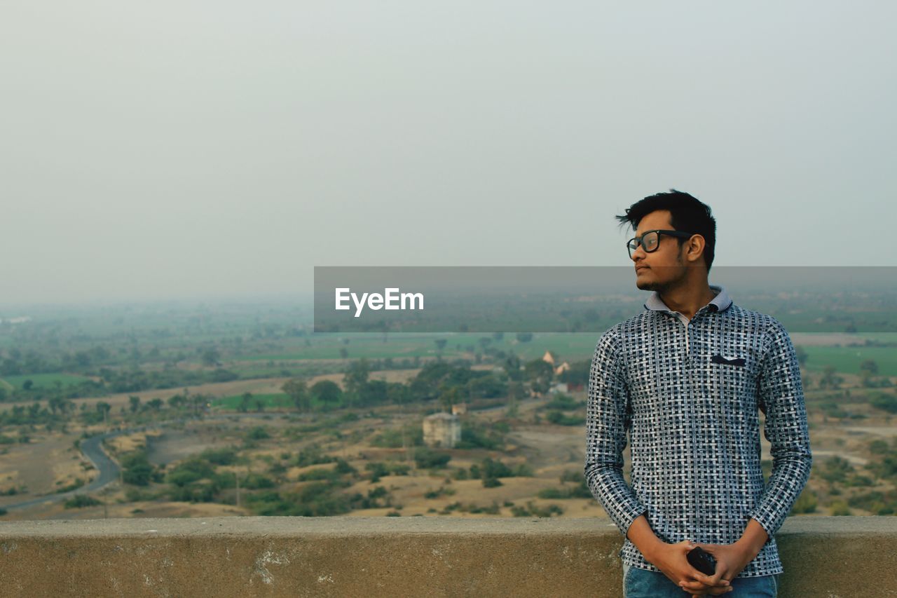 Man looking away while leaning on retaining wall against sky