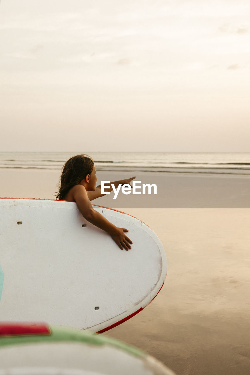 Girl with paddleboard pointing at sea on beach during sunset