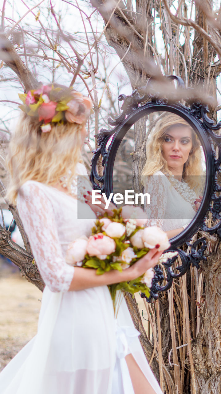 Young woman looking into mirror against bare tree