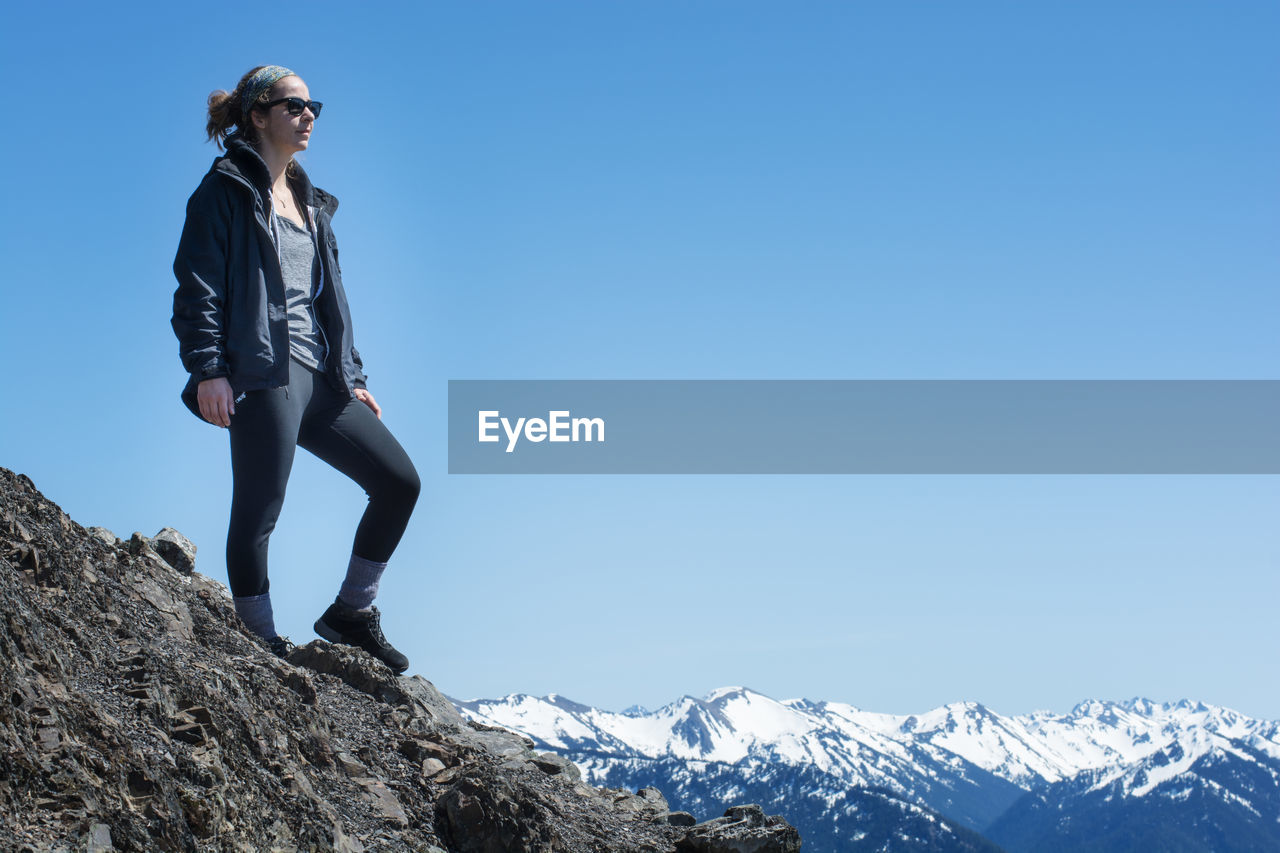 Low angle view of young woman standing on cliff against clear blue sky