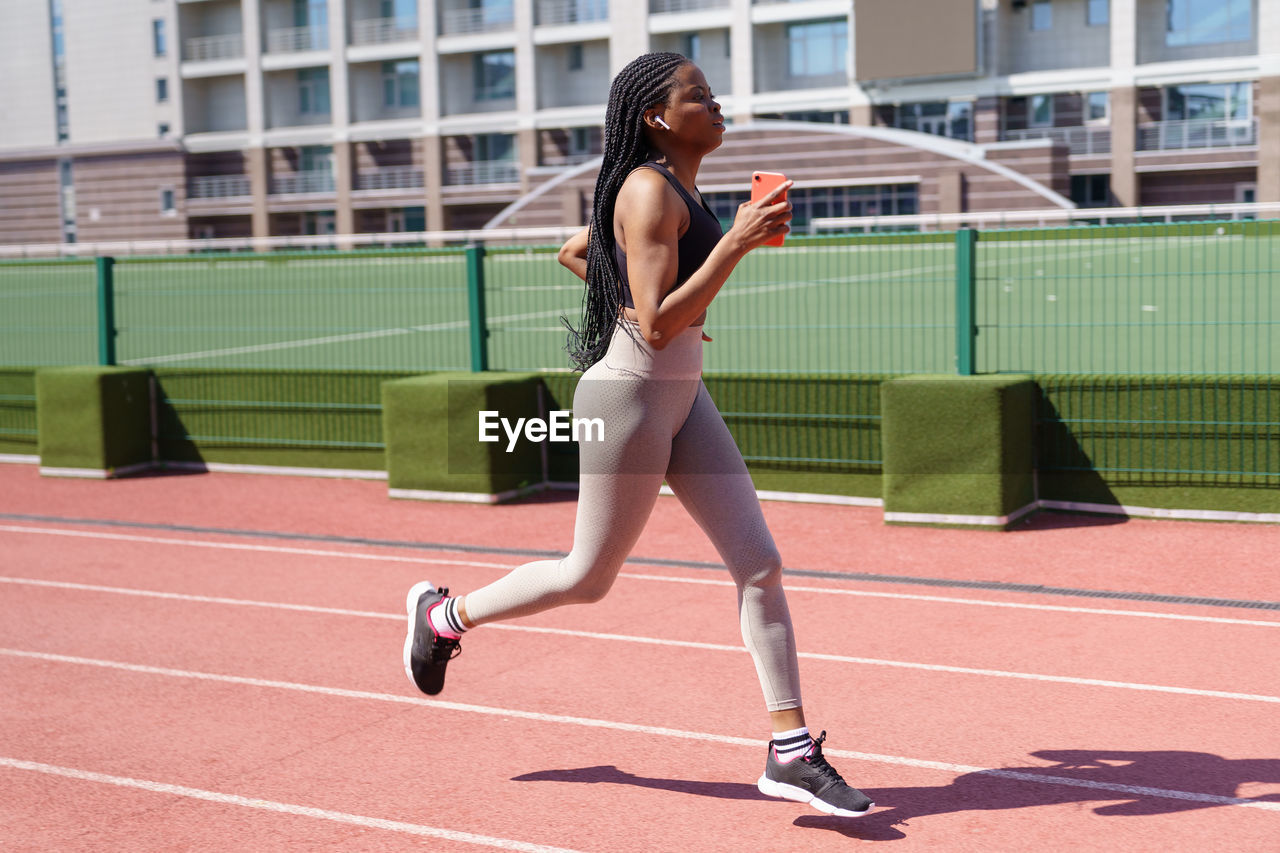 Young woman in headphones runs every day on red track at sports ground of big residential complex