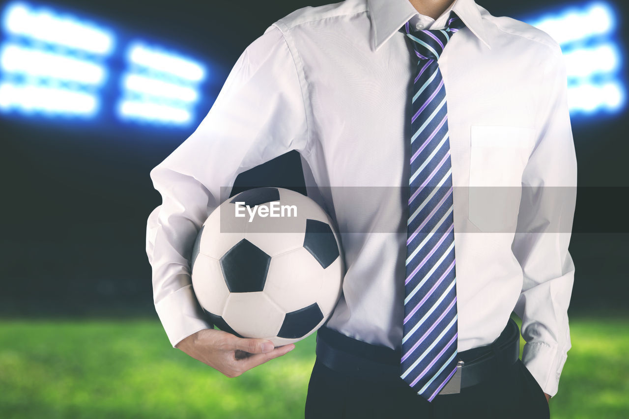 Midsection of businessman holding soccer ball on field at night