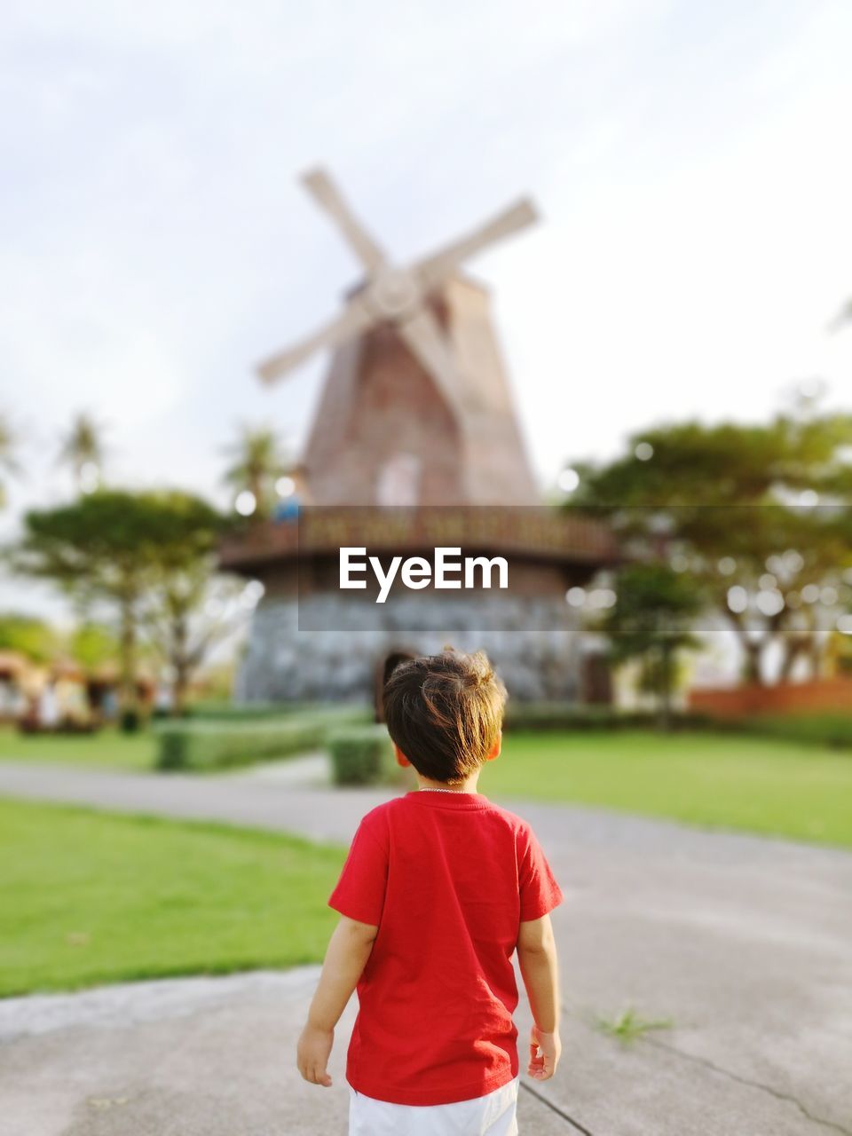 Rear view of boy looking at traditional windmill while standing on footpath against sky