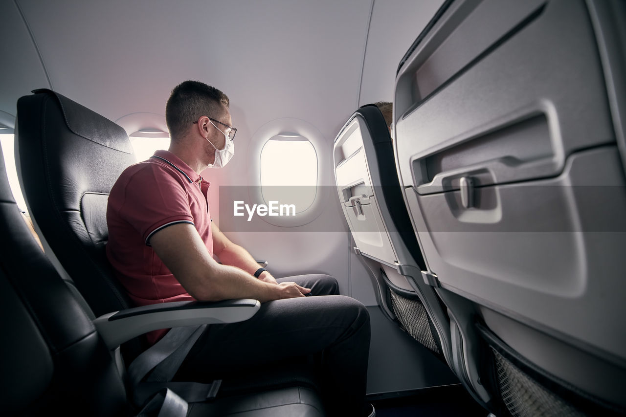 Passenger with protective face mask in airplane looking through window. 