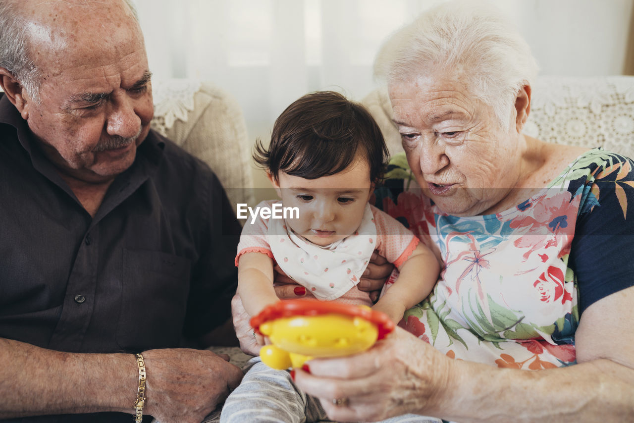 Great-grandparents sitting with baby girl on the couch at home looking at toy