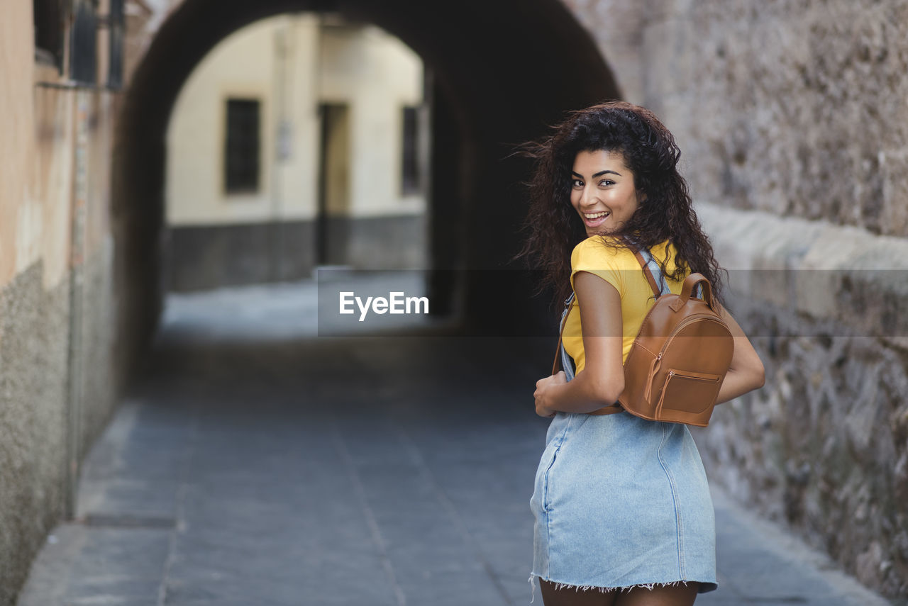 Portrait of smiling young backpack woman standing on street in city