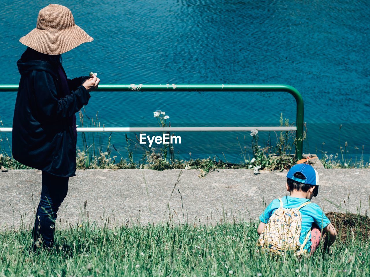 Side view of mother standing by son crouching on grassy field by lake