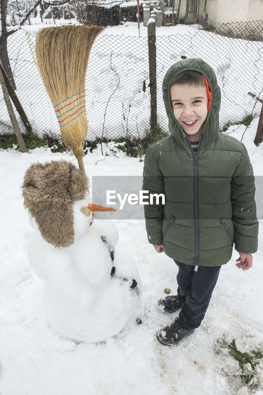 Full length of smiling boy standing by snowman in yard during winter