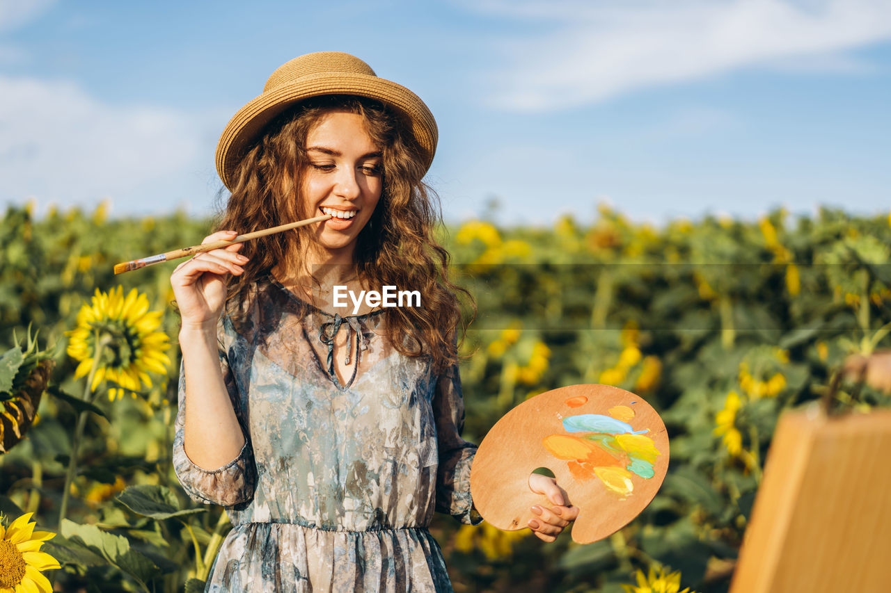 A young woman with curly hair and wearing a hat is painting in nature. 