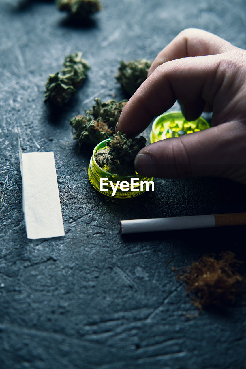 From above faceless man grinding dry cannabis bud while rolling blunt on grunge surface at home