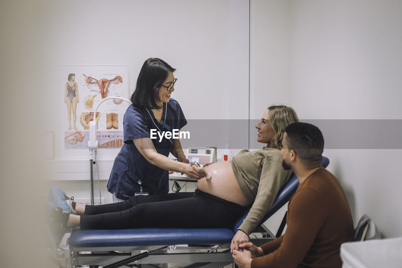 Smiling female doctor doing ultrasound of pregnant woman sitting on examination table by man in hospital