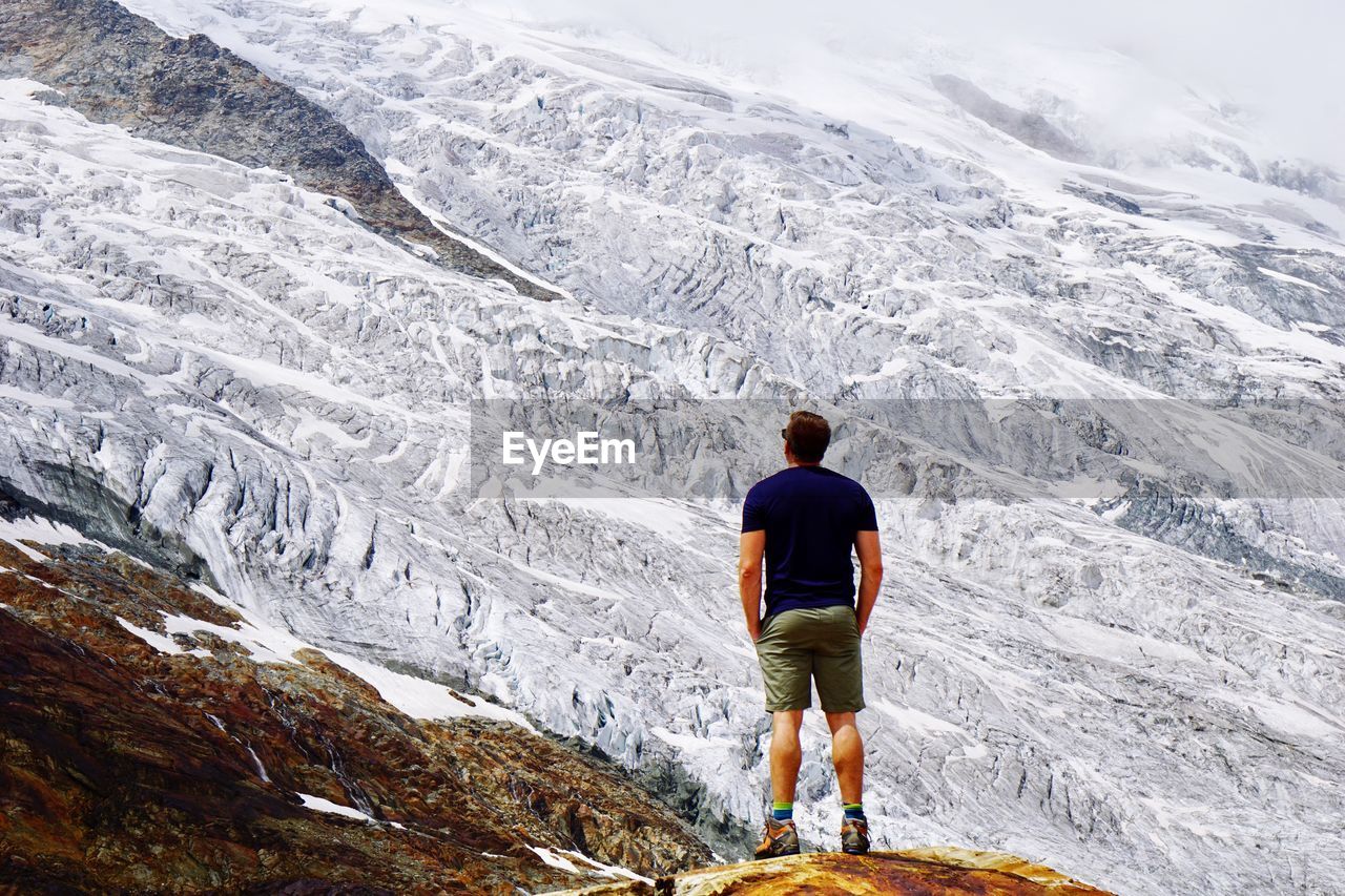 REAR VIEW OF MAN STANDING ON SNOWCAPPED MOUNTAINS