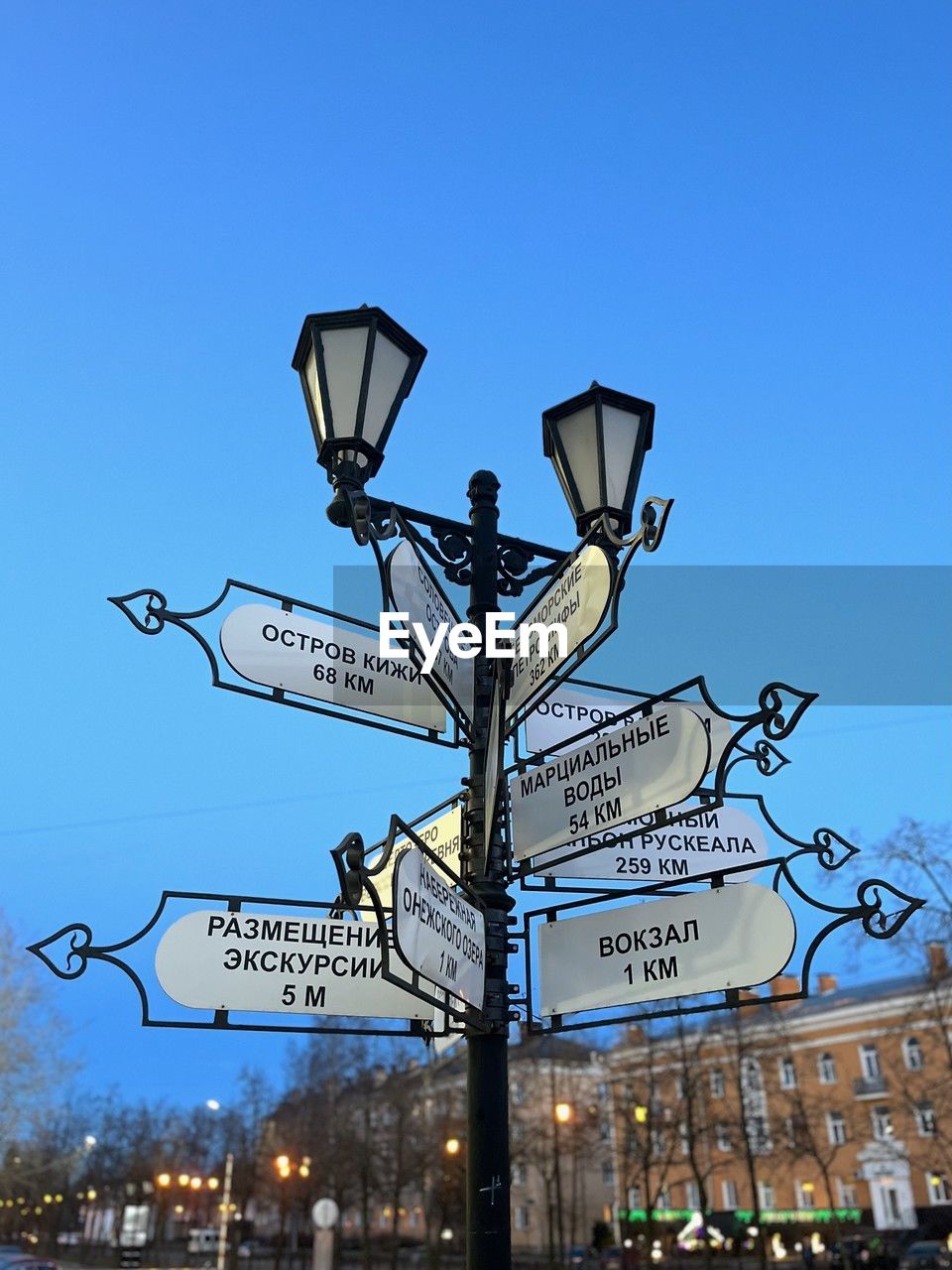 lighting, street light, sign, street, city, sky, communication, light fixture, architecture, illuminated, road, text, nature, guidance, clear sky, blue, lighting equipment, road sign, western script, arrow symbol, transportation, directional sign, light, outdoors, no people, building exterior, built structure, landmark, symbol, day, travel destinations, signage, travel, low angle view, city life, information sign