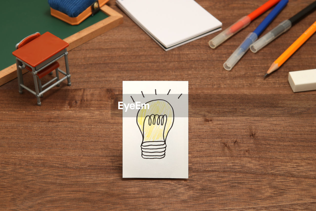 High angle view of light bulb on paper with school supply toys on wooden table