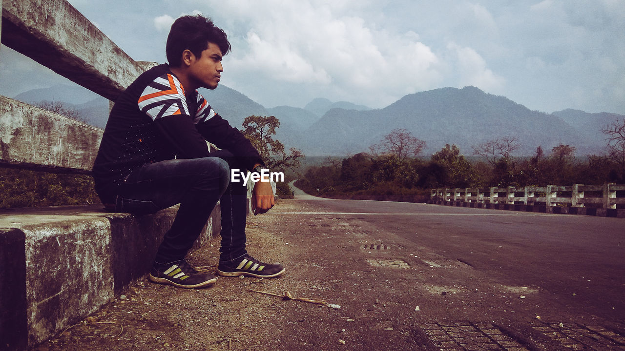 Side view of young man looking away while sitting on road against sky