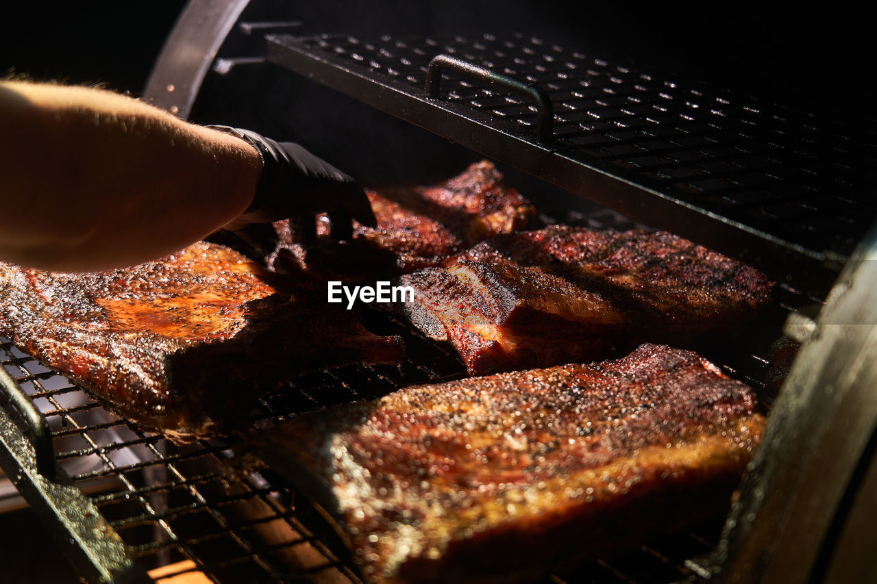 From above of crop chef in gloves grilling chunks of meat in on rack in barbecue