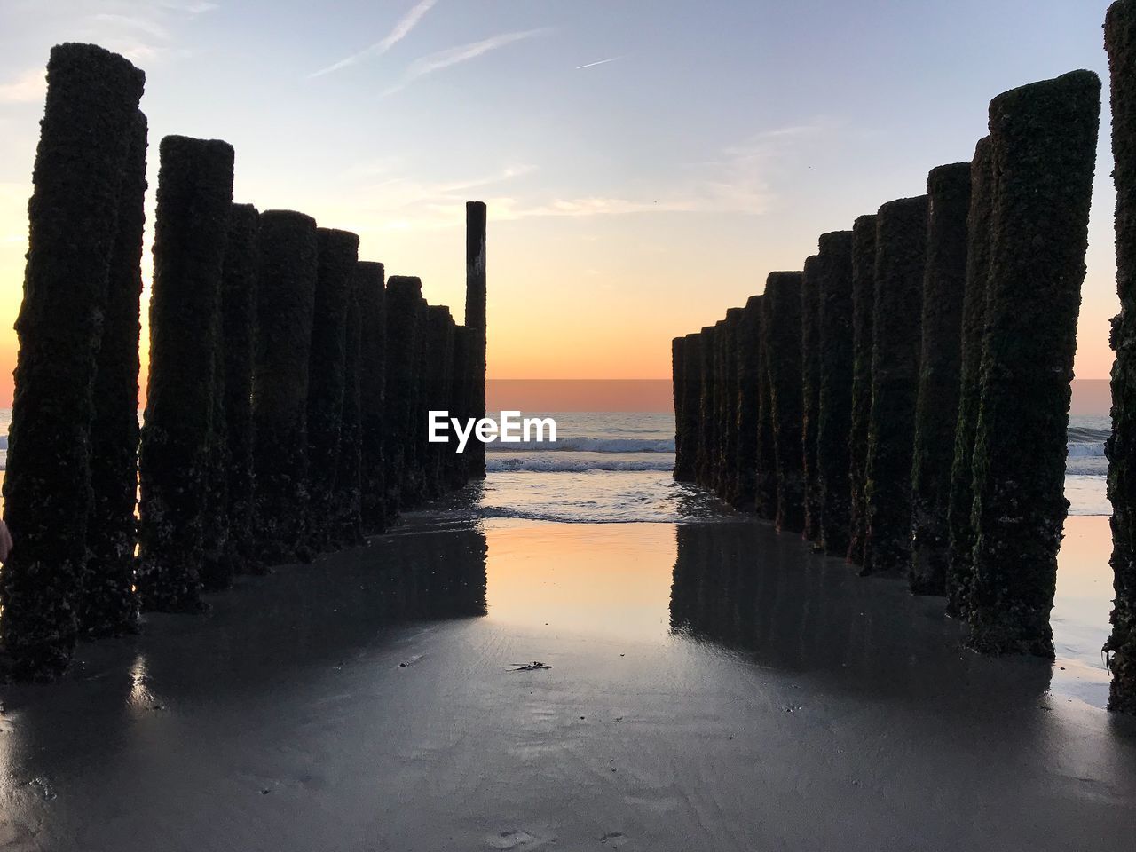 WOODEN POSTS ON SEA AGAINST SKY AT SUNSET