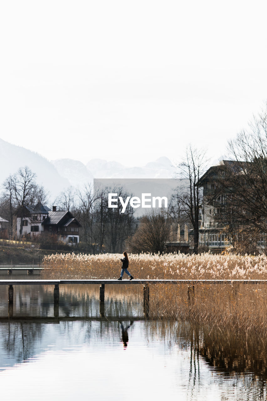 Side view of unrecognizable person walking on wooden boardwalk over calm lake with grassy shore in small settlement located in mountainous terrain in autumn day