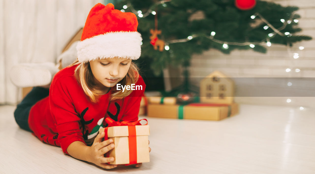 A blonde girl lies on the floor next to a decorated christmas tree and holds a gift tied