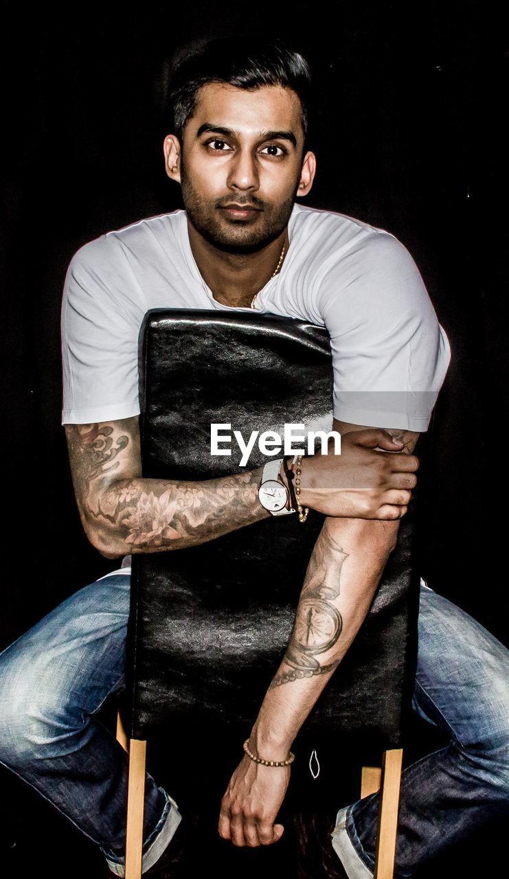 Portrait of young man sitting on chair against black background