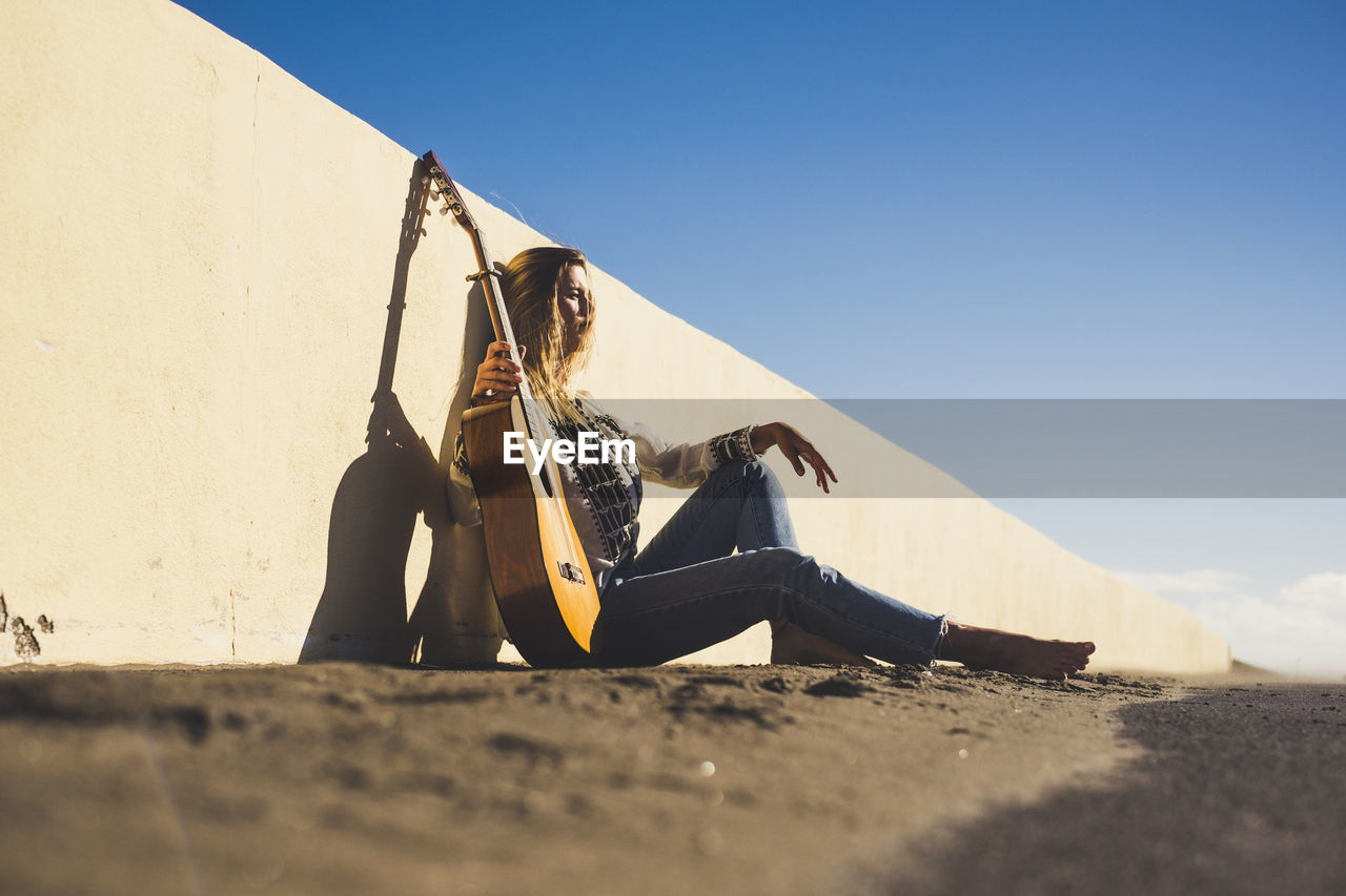 Young woman with guitar sitting on road by wall against clear sky