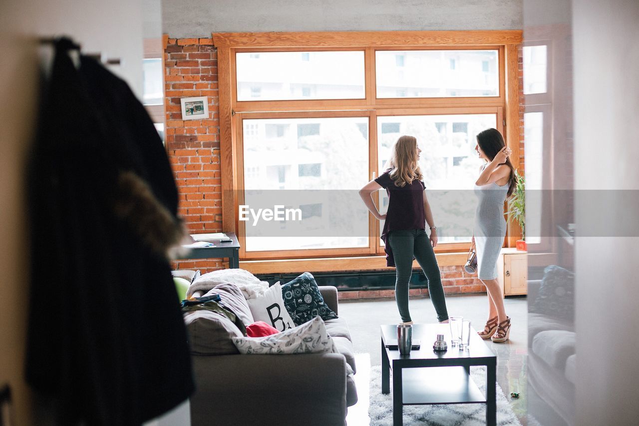 Full length of friends talking while standing by window in living room