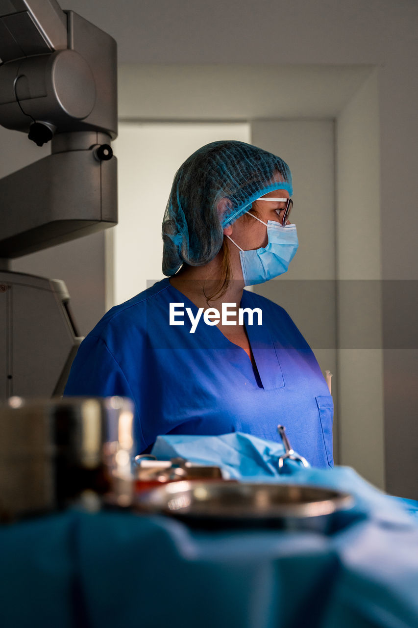 Pensive female doctor in medical uniform looking away while standing against blurred interior in laboratory of contemporary hospital