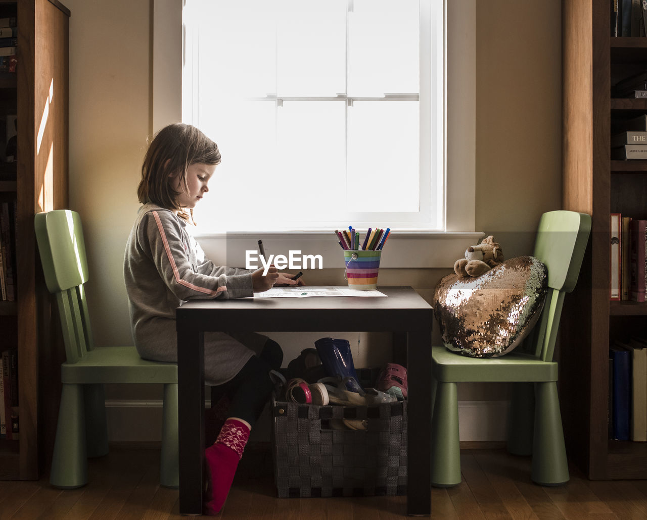 Side view of girl drawing on paper at table against window in home