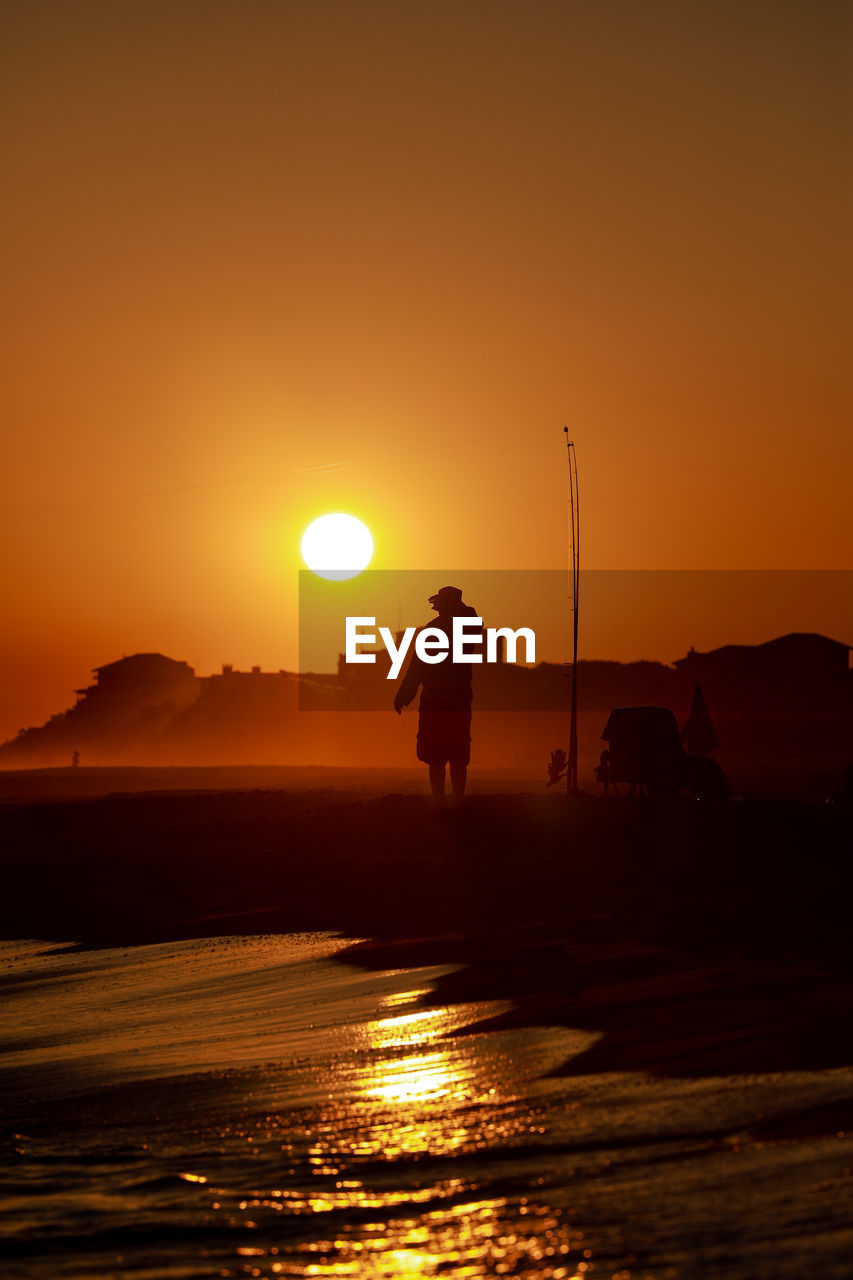 Silhouette man standing at beach against clear orange sky during sunset