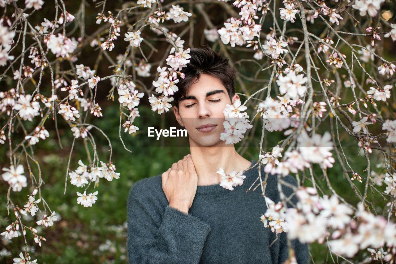 Young male with closed eyes standing under branches with flowers of blooming almond tree in spring garden