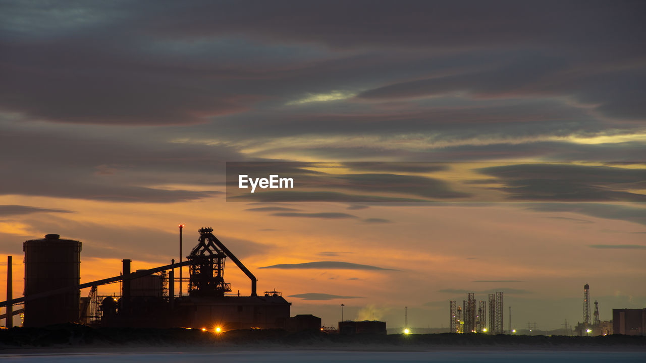 LOW ANGLE VIEW OF ILLUMINATED CRANES AGAINST SKY DURING SUNSET