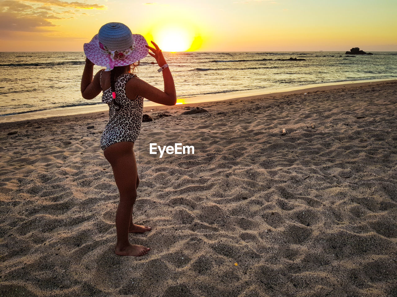 Side view of girl wearing hat standing at beach during sunset