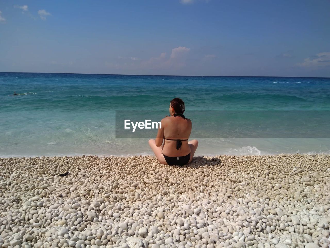 REAR VIEW OF SHIRTLESS MAN SITTING ON BEACH AGAINST SEA