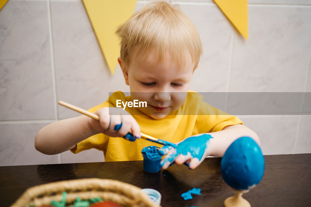 Little boy draws blue paint on his hand, paints an easter egg