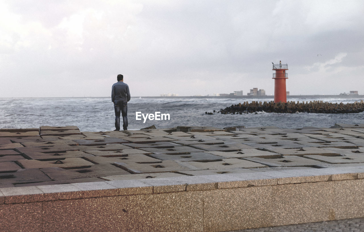 Rear view of man standing on beach against cloudy sky