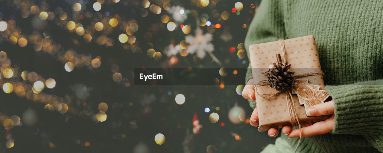 Small gift in woman's hands. close up. decorated christmas tree on background. 