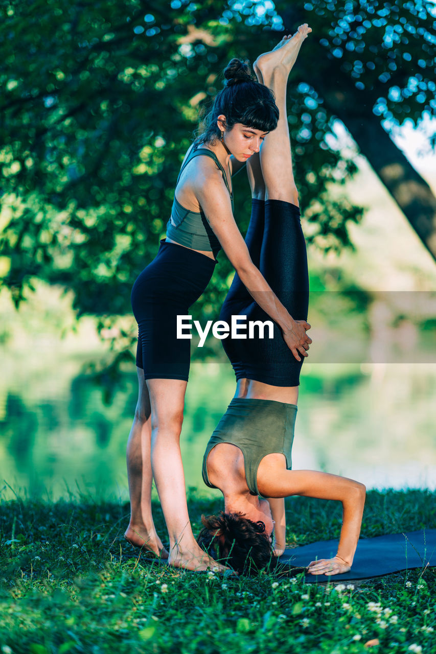 Women doing yoga by the water. instructor helping woman to do headstand.