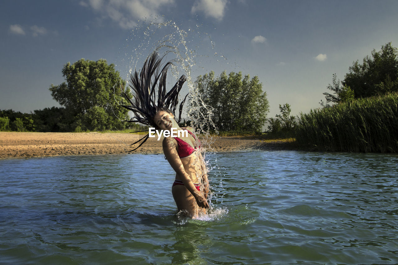 Girl splashing water while tossing hair in lake against sky