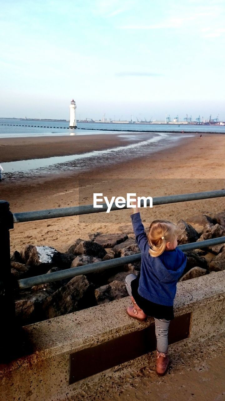 Rear view of girl climbing on railing by sea against sky